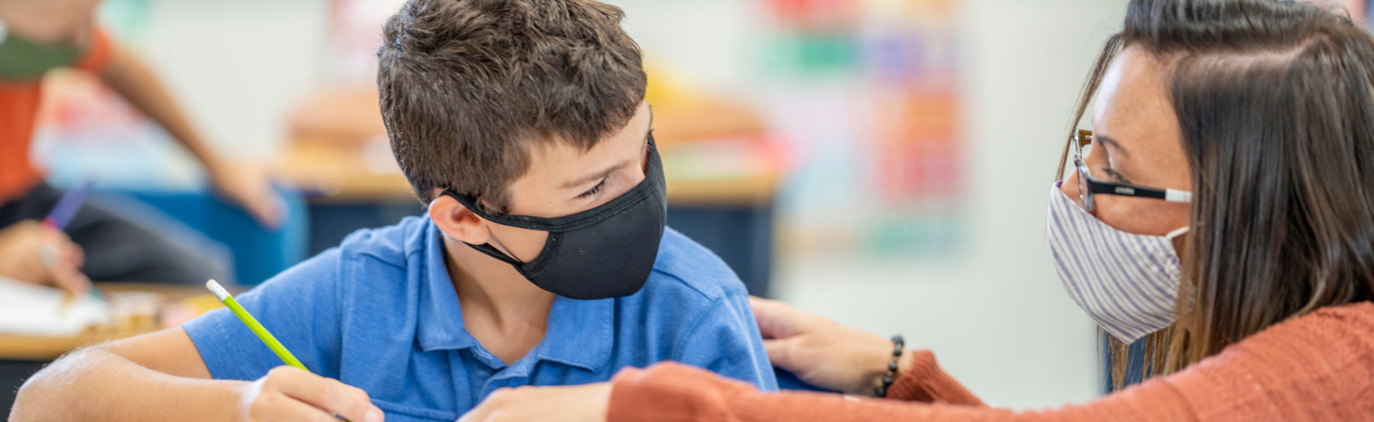 Elementary school teacher helping student, both wearing masks