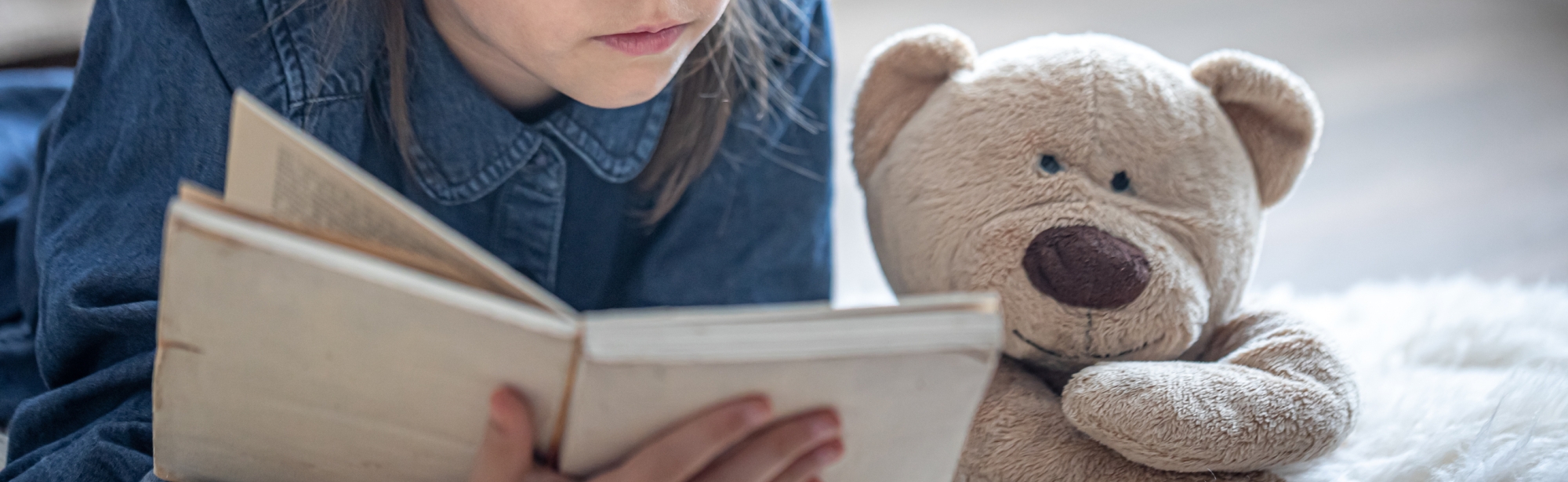 Little girl lying on the floor reads a book next to her teddy bear