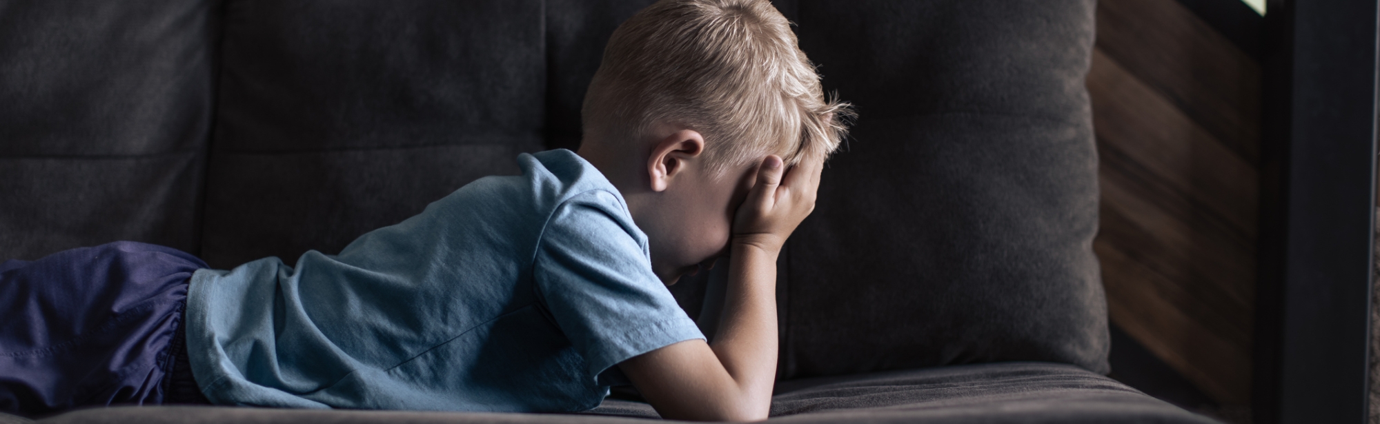 Young boy laying on couch alone, crying with hands over face