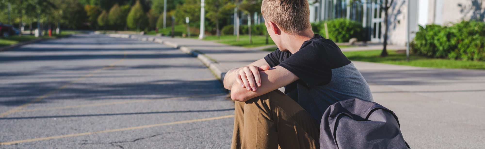 Teen sitting alone on curb of street with backpack